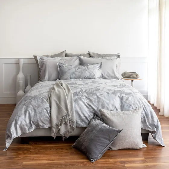 A bed with gray and blue Opus Jacquard Duvet Cover and a selection of gray throw pillows on a wooden floor, with a white wall and a small lamp beside the bed.