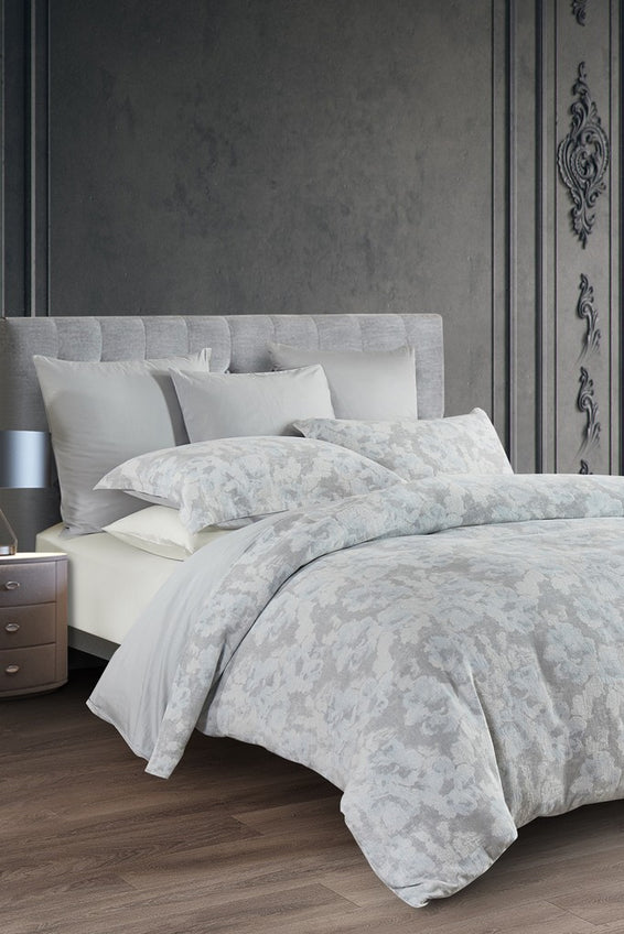 A Mirage duvet cover set with floral tapestry design in greys, warm whites, and soft blue displayed on a bed.