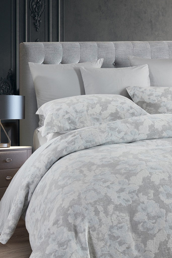 A Mirage duvet cover set with floral tapestry design in greys, warm whites, and soft blue displayed on a bed.