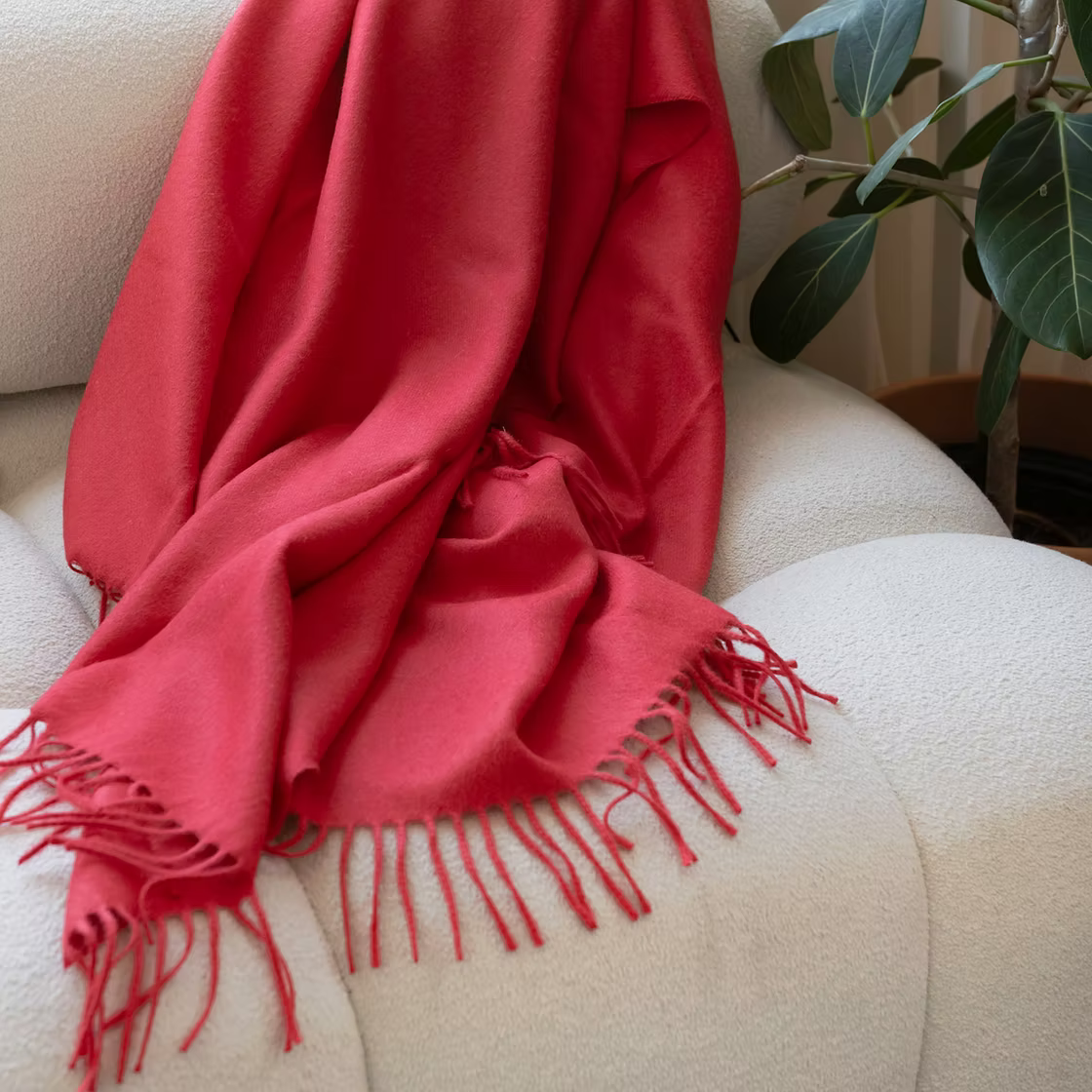 Red Alfie Baby Alpaca Throw iwith fringes draped over a beige armchair, with a plant in the background.