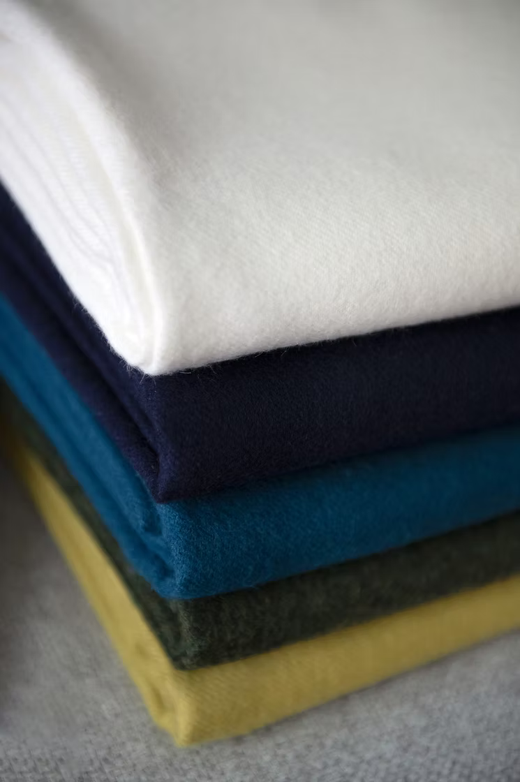 Stack of Alfie Baby Alpaca Throws in white, navy, blue, green, and yellow on a textured surface.
