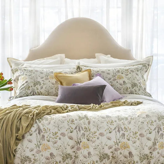 A bed with floral print bedding Mirabel, featuring a variety of colors including beige, green, and purple, with a light background wall and a beige headboard.