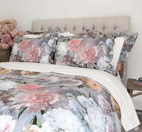 A bed with a floral Bijoux Duvet Cover featuring pink and white flowers, placed in a bedroom setting with decorative pillows and a nightstand in the background.