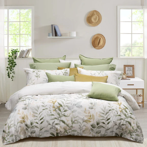 A set of Aspen Duvet Cover Set with a botanical design in green and gold tones, displayed on a bed in a room with a white shelf, a straw hat, and a small wooden table.