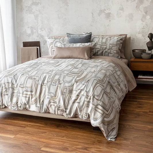 A bed with Fjord Jacquard bedding in a geometric pattern of blues and taupes, placed in a room with wooden flooring and a textured wall.