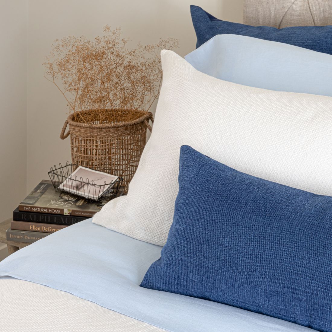 A bed with blue and white linens, including Avalon Honeycomb Linen Corvelet, pillows, and a basket with dried plants in the background.