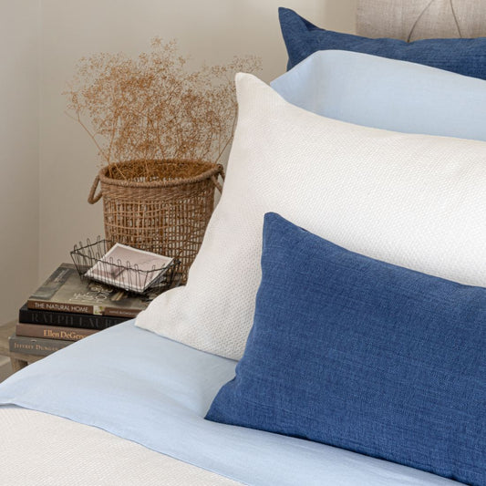 A set of blue and white linen Avalon Honeycomb Linen Cushions on a bed, with a decorative item and books on the side table.